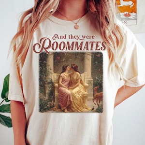 May include: Ivory-colored t-shirt featuring the text "And they were Roommates" above a vintage-style image of two women kissing. The image is framed within a rectangle. The shirt is a neutral color.