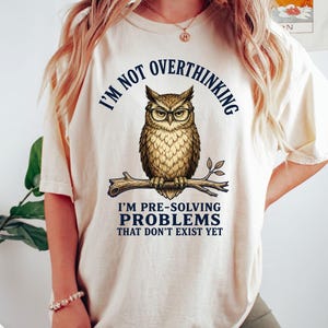 Pode incluir: Uma t-shirt de manga curta na cor marfim com o gráfico de uma coruja castanha de óculos pousada num ramo. O texto diz I'M NOT OVERTHINKING I'M PRE-SOLVING PROBLEMS THAT DON'T EXIST YET em letras azul-escuras acima e abaixo da coruja.