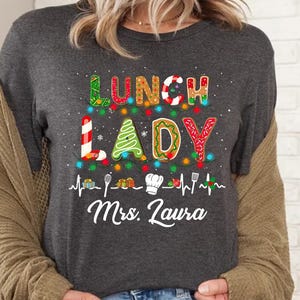 May include: Dark grey heather t-shirt with the words "LUNCH LADY" in colorful, festive lettering. The design includes a Christmas tree, candy cane, and chef's hat, with the name "Mrs. Laura" below. A fun, holiday-themed shirt.