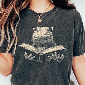 May include: Dark grey t-shirt with a beige illustration of a bearded dragon wearing glasses and reading a book. The word "PEPPER" is printed on a black circle at the bottom left. The shirt is a casual style, suitable for everyday wear.