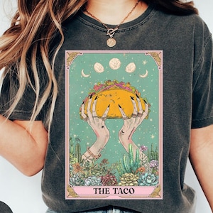 May include: Dark grey t-shirt featuring a tarot card design with a taco held by hands. The card has a teal background with moons and cacti. The text "THE TACO" is at the bottom of the card.