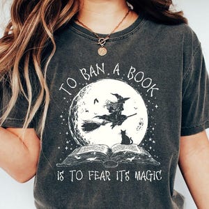 以下が含まれることがあります： 「TO BAN A BOOK IS TO FEAR ITS MAGIC」の文字が入った濃いグレーのTシャツ。満月の前をほうきに乗った魔女、黒猫、開いた本が描かれたグラフィック。シャツの色はペッパーです。