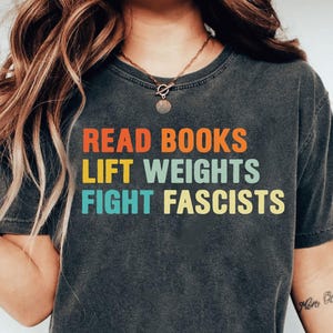 Könnte beinhalten: Dunkelgraues T-Shirt mit dem Text "READ BOOKS LIFT WEIGHTS FIGHT FASCISTS" in Orange, Gelb und Türkis. Das Shirt hat einen Rundhalsausschnitt und kurze Ärmel. Die Person trägt eine blaue Jeans.