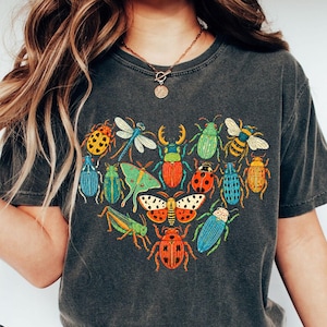Op de afbeelding: Een antracietgrijs t-shirt met een hartvormige afbeelding bestaande uit diverse kleurrijke insecten, waaronder kevers, een libel, een mot en een sprinkhaan. Het t-shirt heeft korte mouwen en een ronde hals, met het woord PEPPER eronder gedrukt.