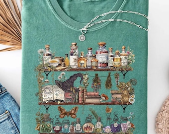 Puede incluir: Una camiseta verde azulado con un diseño gráfico detallado de botellas de pociones, libros y un sombrero de bruja en estanterías de madera. Un collar plateado con un colgante de sol descansa sobre la camiseta. La camiseta está doblada, con unos vaqueros azules y un zapato visibles.