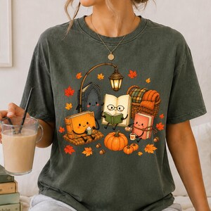 Puede incluir: Una camiseta gris oscuro de manga corta con un estampado gráfico de tres libros antropomórficos con caras, una farola brillante, una calabaza y hojas de otoño naranjas dispersas. Los libros aparecen leyendo, bebiendo y relajándose.