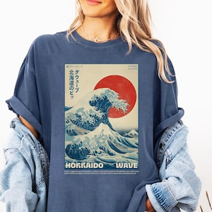 May include: Denim blue t-shirt featuring a Japanese-inspired wave design with a red sun and the words "Hokkaido Wave". The graphic includes Japanese text. The shirt is a relaxed fit.