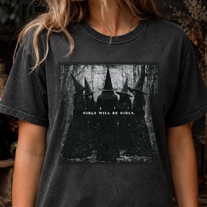 Può includere: Una t-shirt a maniche corte grigio antracite con una stampa grafica rettangolare di cinque figure che indossano cappelli a punta in un ambiente boschivo. Il testo "GIRLS WILL BE GIRLS." è stampato in bianco al centro del disegno.