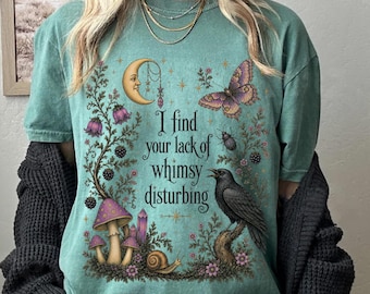 May include: A light green short-sleeve t-shirt featuring a graphic print of a crescent moon, a butterfly, a crow, mushrooms, and a snail. The text "I find your lack of whimsy disturbing" is printed in the center. The shirt is worn with a dark cardigan.