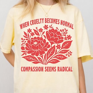 Op de afbeelding: Lichtgele t-shirt met een rood bloemenontwerp en de tekst "WHEN CRUELTY BECOMES NORMAL COMPASSION SEEMS RADICAL". Het shirt heeft korte mouwen en een ronde hals.