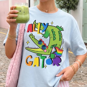 May include: A light blue t-shirt featuring a graphic of a green alligator wearing heart-shaped rainbow glasses. The text "ALLY GATOR" is printed in colorful, bold letters above and below the alligator, which holds a small pride flag.