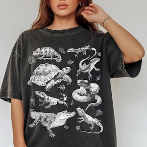 May include: Dark grey t-shirt with a white illustration of reptiles, such as a turtle, snake, and alligator. The design is printed on a vintage-washed, oversized tee, giving a relaxed, casual look. The shirt has short sleeves and a crew neck.