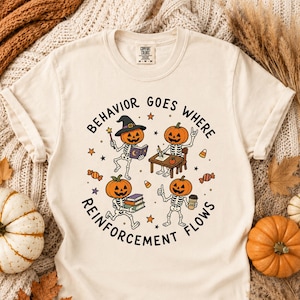 Op de afbeelding: Een crèmekleurig T-shirt met korte mouwen en een afbeelding van vier skeletten met pompoenhoofden. De tekst luidt BEHAVIOR GOES WHERE REINFORCEMENT FLOWS. De skeletten zijn afgebeeld terwijl ze lezen, aan een bureau schrijven, boeken dragen en een koffiebeker vasthouden.