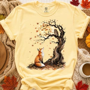 Könnte beinhalten: Ein blassgelbes T-Shirt mit einem Grafikdruck eines Fuchses, der unter einem knorrigen Baum sitzt, auf dessen Ast eine Eule thront. Die Szene zeigt einen großen Vollmond und fallende Herbstblätter. Das Shirt hat einen Rundhalsausschnitt.