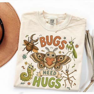 May include: A cream-coloured t-shirt featuring a graphic print of various cartoon insects, including a beetle, a moth holding a heart, a praying mantis, a caterpillar, and a stick insect, with the text "BUGS NEED HUGS" in orange and green.