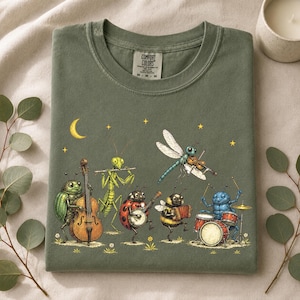 Op de afbeelding: Een olijfgroen t-shirt met een grafische print van insecten die muziekinstrumenten bespelen, waaronder een kever met een contrabas, een bidsprinkhaan met een fluit, een lieveheersbeestje met een banjo, een libel met een viool, een bij met een accordeon en een blauwe kever met drums.