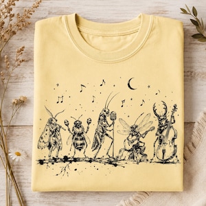 May include: A pale yellow crew neck t-shirt featuring a black ink illustration of five insects playing musical instruments. The graphic depicts a moth with a flute, a bee with maracas, a cricket at a microphone, a dragonfly with a guitar, and a beetle with a double bass, surrounded by musical notes and a crescent moon.