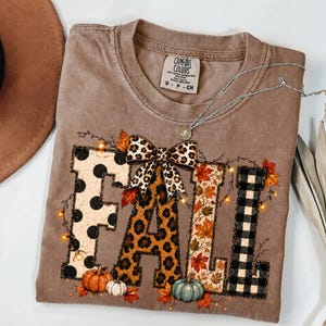 May include: A tan short-sleeved t-shirt featuring the word FALL in large block letters. Each letter has a different pattern: polka dots, leopard print, autumn leaves, and black plaid. A leopard print bow sits above the letters with pumpkins.