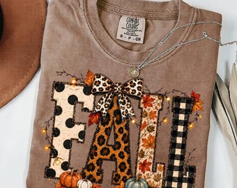 Puede incluir: Una camiseta de manga corta color canela con la palabra FALL en letras grandes. Cada letra tiene un patrón diferente: lunares, estampado de leopardo, hojas de otoño y cuadros negros. Un lazo de leopardo se sitúa sobre las letras.