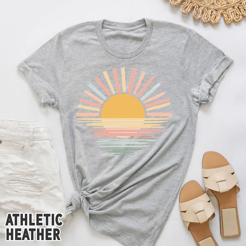 May include: Athletic heather gray t-shirt featuring a colorful sunset design. The graphic includes a yellow sun, pastel rays, and a water reflection in shades of pink, orange, and green. The shirt is paired with white shorts and beige sandals.