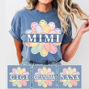 以下が含まれることがあります： パステルカラーの生地パッチで作られた花の模様が特徴の青いTシャツ。「MIMI」の文字が中央に表示され、その下に「freya noah olivia」の名前があります。「おばあちゃん」と「ナナ」の追加デザインも表示されています。