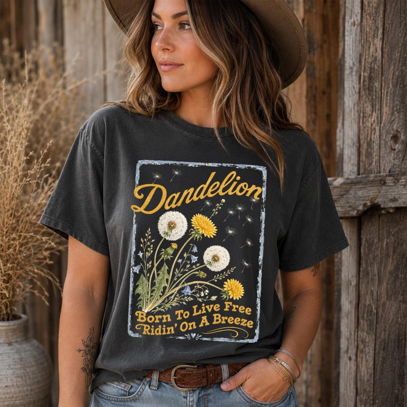 May include: Dark gray t-shirt with a dandelion graphic. The graphic features the word "Dandelion" in yellow script, along with illustrations of dandelions and the text "Born To Live Free Ridin' On A Breeze."
