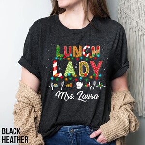 May include: Black heather t-shirt with the words "LUNCH LADY" in colorful, festive lettering. The design includes a Christmas tree, candy cane, and sprinkles, with a heartbeat line and the name "Mrs. Laura" below. The shirt is a crew neck.