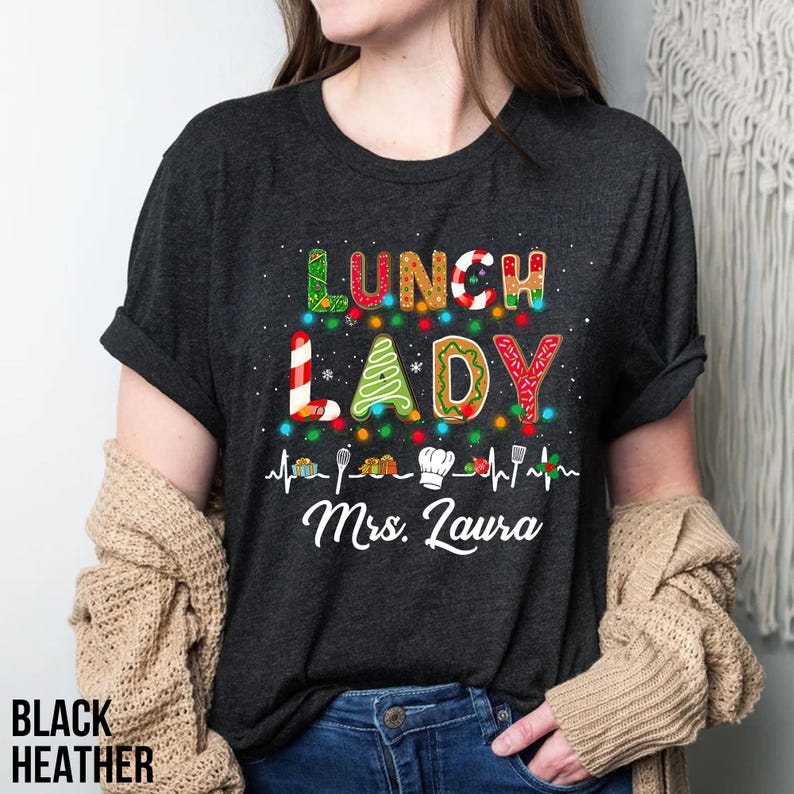 May include: Black heather t-shirt with the words "LUNCH LADY" in colorful, festive lettering. The design includes a Christmas tree, candy cane, and sprinkles, with a heartbeat line and the name "Mrs. Laura" below. The shirt is a crew neck.