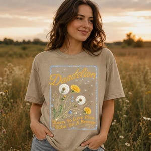 May include: A tan t-shirt featuring a dandelion design with the word "Dandelion" in yellow script. The design includes white and yellow dandelions, blue flowers, and the text "Born To Live Free Ridin' On A Breeze."