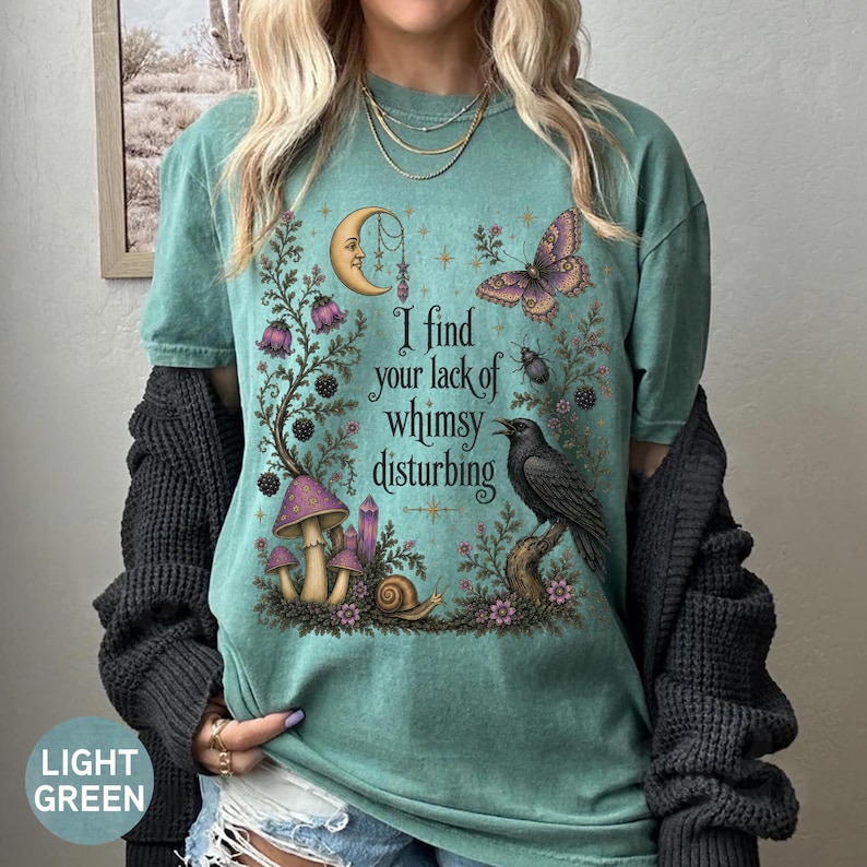 Pode incluir: Uma t-shirt de manga curta verde-clara com uma impress&atilde;o gr&aacute;fica de uma lua crescente, uma borboleta, um corvo, cogumelos e um caracol. O texto "I find your lack of whimsy disturbing" est&aacute; impresso no centro da t-shirt.