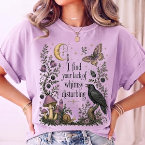 Pode incluir: Uma t-shirt roxo-claro com uma estampa gr&aacute;fica com o texto I find your lack of whimsy disturbing. O design inclui uma lua crescente, uma borboleta, um corvo preto, cogumelos, um caracol e elementos bot&acirc;nicos em tons de roxo escuro.