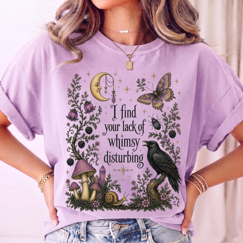 Pode incluir: Uma t-shirt roxo-claro com uma estampa gr&aacute;fica com o texto I find your lack of whimsy disturbing. O design inclui uma lua crescente, uma borboleta, um corvo preto, cogumelos, um caracol e elementos bot&acirc;nicos em tons de roxo escuro.