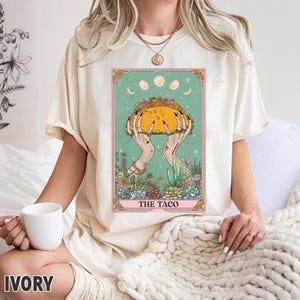 May include: Ivory-colored t-shirt featuring a tarot card design with a taco held by hands. The card has a teal background with moon phases and floral elements. The text "THE TACO" is at the bottom.