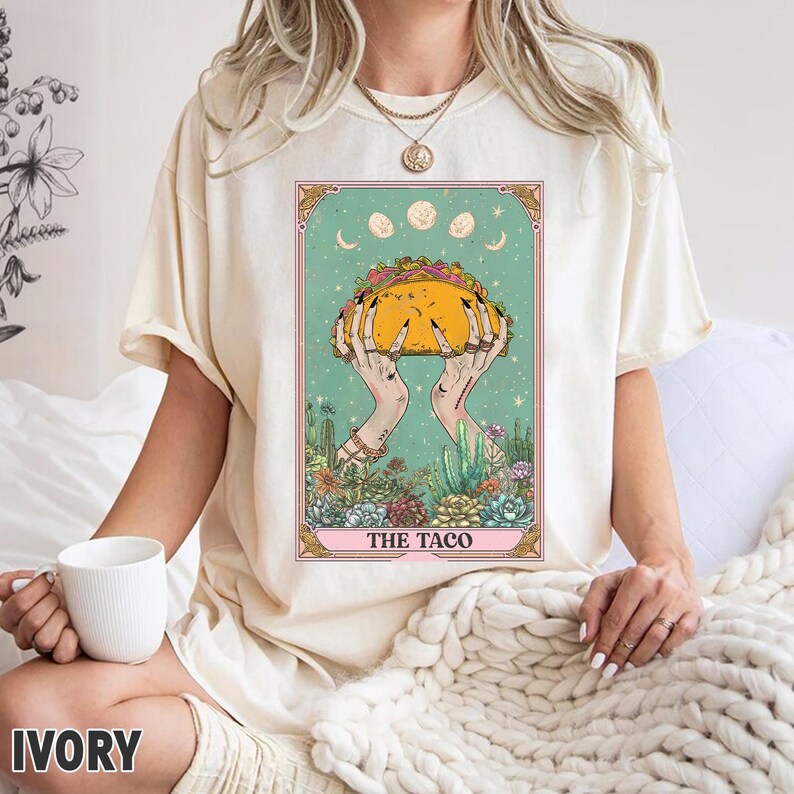 May include: Ivory-colored t-shirt featuring a tarot card design with a taco held by hands. The card has a teal background with moon phases and floral elements. The text "THE TACO" is at the bottom.