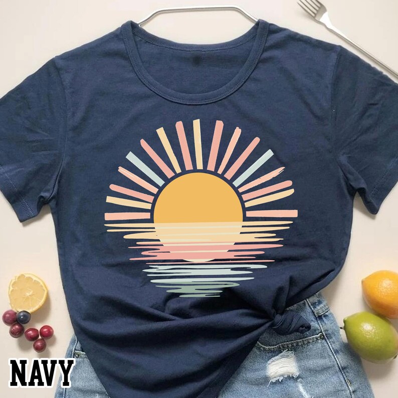 May include: Navy blue t-shirt featuring a colorful sunset design. The graphic includes a yellow sun, pastel rays, and a reflection on the water. The word "NAVY" is printed at the bottom left corner of the shirt.