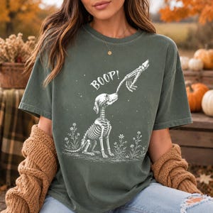 以下が含まれることがあります： 暗いオリーブグリーンの半袖Tシャツ。花の中に座る骸骨の犬の白いグラフィックプリントが施されています。骸骨の指が犬の鼻に触れており、犬の頭上には白い文字でBOOPとプリントされています。