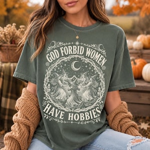 Puede incluir: Una camiseta de manga corta color verde oliva oscuro con un estampado gráfico blanco de cuatro brujas con sombreros bailando en círculo bajo una luna creciente. El texto "GOD FORBID WOMEN HAVE HOBBIES" aparece en una fuente curva alrededor.