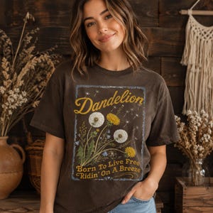 May include: Brown t-shirt with a dandelion graphic. The design features the word "Dandelion" in yellow script above an illustration of dandelions and the text "Born To Live Free Ridin' On A Breeze".