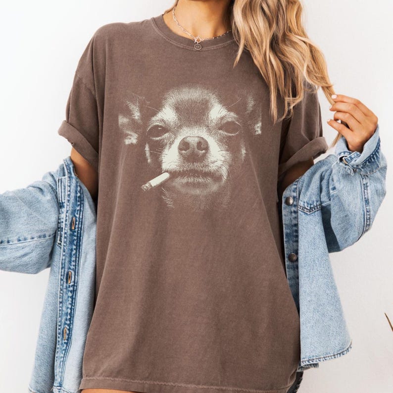 May include: Brown t-shirt with a black and white graphic of a Chihuahua smoking a cigarette. The tee has short sleeves and a relaxed fit. The dog's face is detailed, with a slightly amused expression.