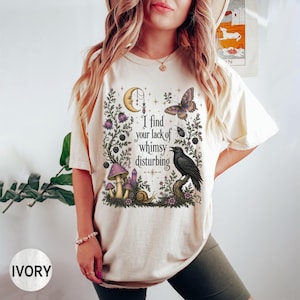 Pode incluir: Uma t-shirt de manga curta na cor marfim com uma estampa gr&aacute;fica de uma lua crescente, uma borboleta, um corvo, cogumelos e um caracol. O texto "I find your lack of whimsy disturbing" est&aacute; impresso ao centro em fonte preta.