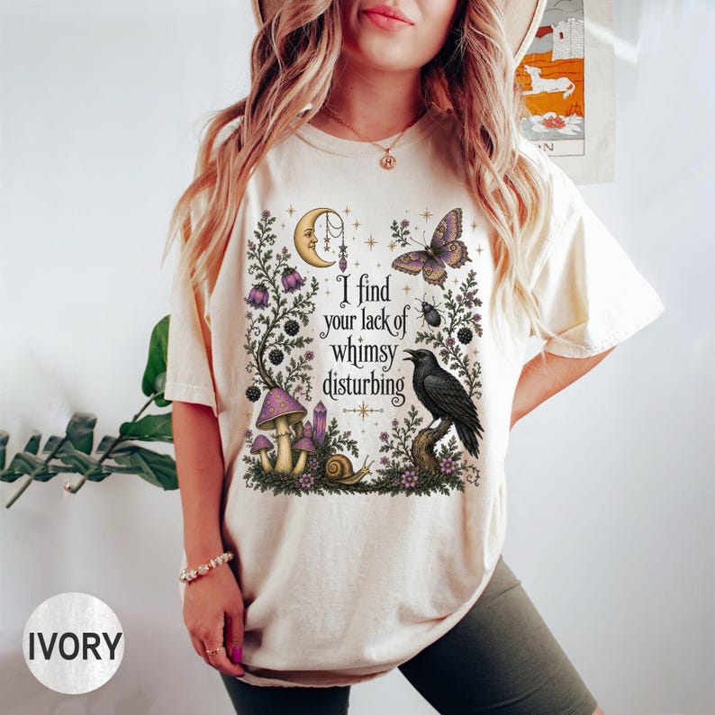 Pode incluir: Uma t-shirt de manga curta na cor marfim com uma estampa gr&aacute;fica de uma lua crescente, uma borboleta, um corvo, cogumelos e um caracol. O texto "I find your lack of whimsy disturbing" est&aacute; impresso ao centro em fonte preta.