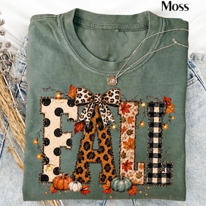 May include: A sage green t-shirt featuring the word FALL in large block letters. Each letter has a different pattern: black polka dots, leopard print, autumn leaves, and black buffalo plaid. A leopard print bow sits atop the letters.