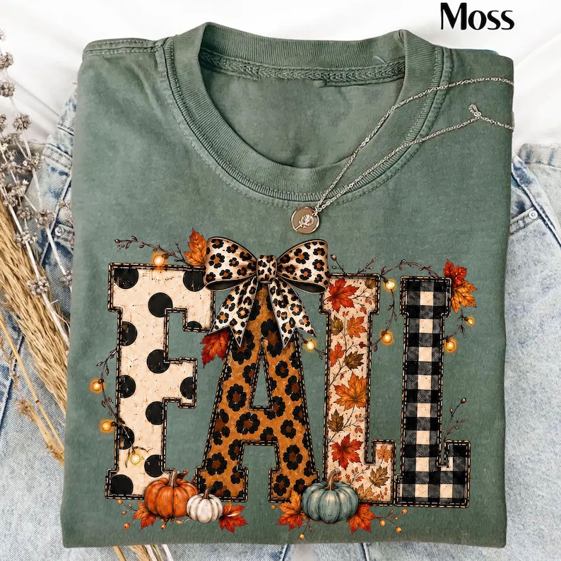 May include: A sage green t-shirt featuring the word FALL in large block letters. Each letter has a different pattern: black polka dots, leopard print, autumn leaves, and black buffalo plaid. A leopard print bow sits atop the letters.