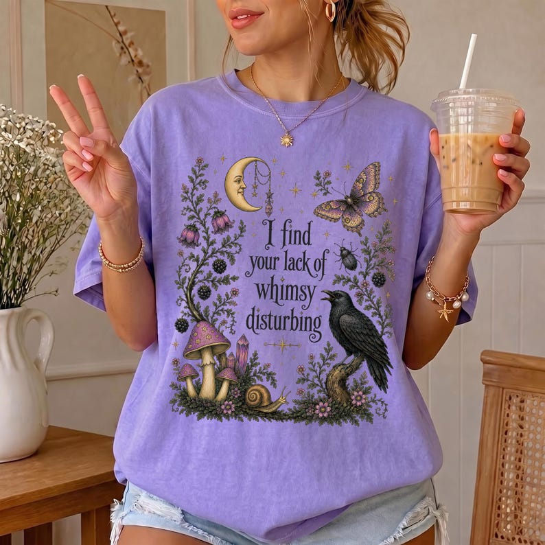 Pode incluir: Uma t-shirt roxo-claro com uma impress&atilde;o gr&aacute;fica de uma lua crescente, uma borboleta, um caracol, cogumelos e um corvo preto. O texto diz, I find your lack of whimsy disturbing. O design inclui elementos florais e estrelas em tons de roxo, amarelo e preto.