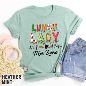 May include: Heather mint green t-shirt with the words "LUNCH LADY" in a festive design with Christmas lights and gingerbread-style letters. Below the text is "Mrs. Laura" in cursive, with cooking-themed graphics.