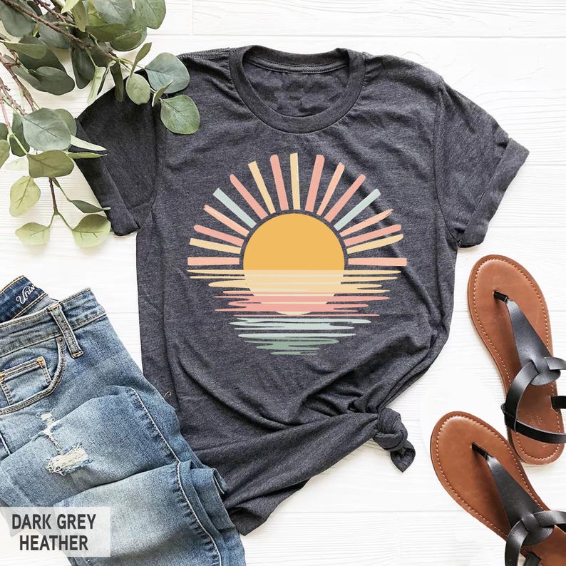 May include: Dark grey heather t-shirt featuring a colorful sunset design. The graphic includes a yellow sun with pastel rays and a reflection on the water. The text "DARK GREY HEATHER" is at the bottom left. The shirt is paired with blue jeans and brown sandals.
