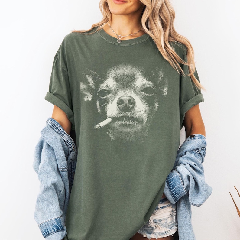 May include: An olive green t-shirt with a black and white image of a Chihuahua smoking a cigarette. The shirt has short sleeves and a relaxed fit. A denim jacket is worn over the shoulders.