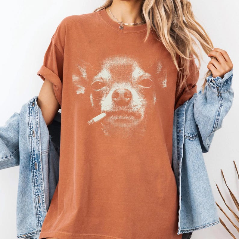 May include: A rust-coloured t-shirt with a detailed graphic of a Chihuahua smoking a cigarette. The dog's face is in close-up, highlighting its expression. The shirt has short sleeves and a relaxed fit, suitable for casual wear.