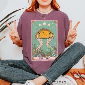 May include: Purple t-shirt featuring a tarot card design with a taco held by hands. The card has a teal background with a crescent moon and three full moons. The text "THE TACO" is at the bottom of the card. The design includes cacti and succulents.