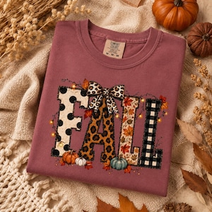 May include: A mauve crewneck t-shirt featuring the word FALL in large block letters. Each letter has a different pattern: black polka dots, leopard print, autumn leaves, and black buffalo plaid. A leopard print bow sits atop the letters.